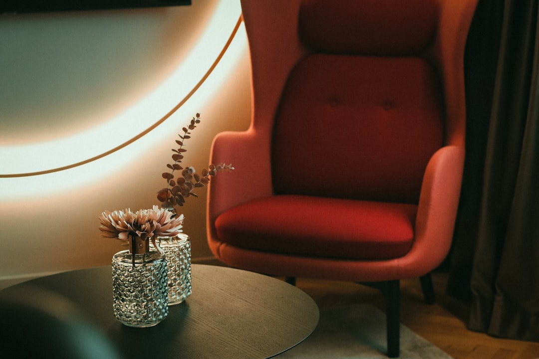 a-red-chair-sitting-next-to-a-table-with-a-vase-of-flowers-djvhh21nueg